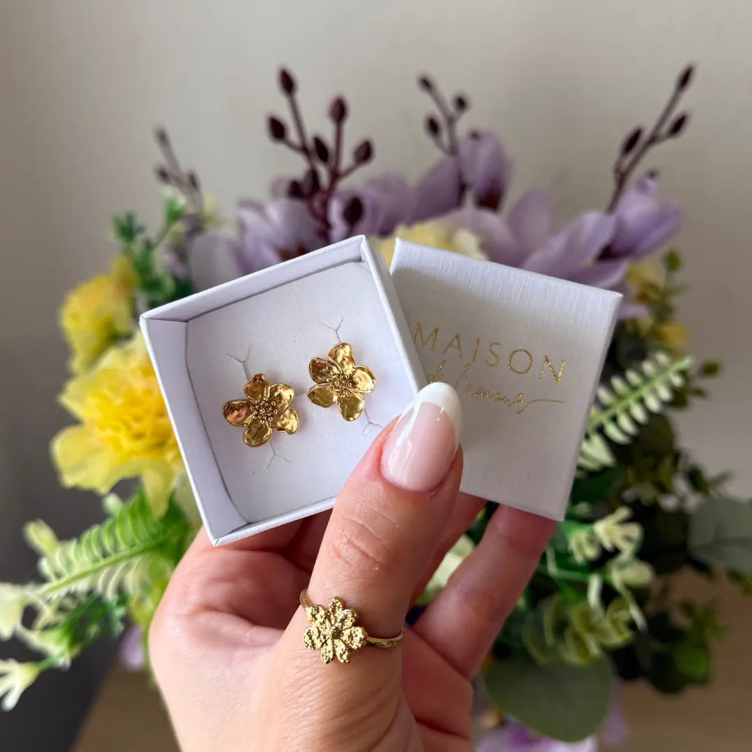Une main tient une boîte blanche ouverte avec deux puces d'oreilles fleurs de cerisier en acier inoxydable doré. Logo Maison