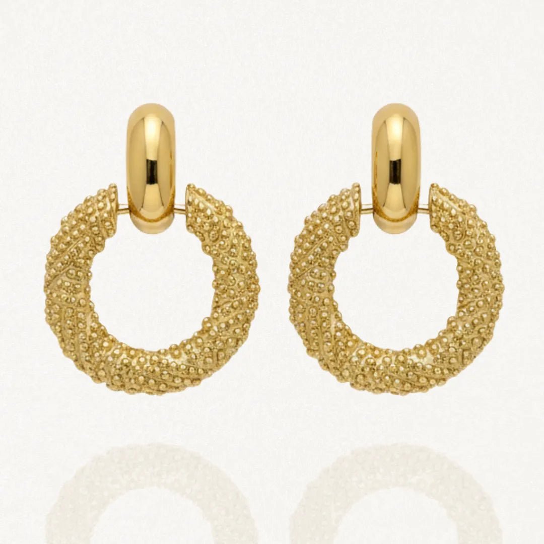 Paire de boucles d'oreilles pendantes dorées, composées d'une créole épaisse avec une texture granulée, suspendue à une attache ovale lisse et brillante. L'image est sur fond blanc avec des reflets.