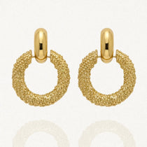 Paire de boucles d'oreilles pendantes dorées, composées d'une créole épaisse avec une texture granulée, suspendue à une attache ovale lisse et brillante. L'image est sur fond blanc avec des reflets.