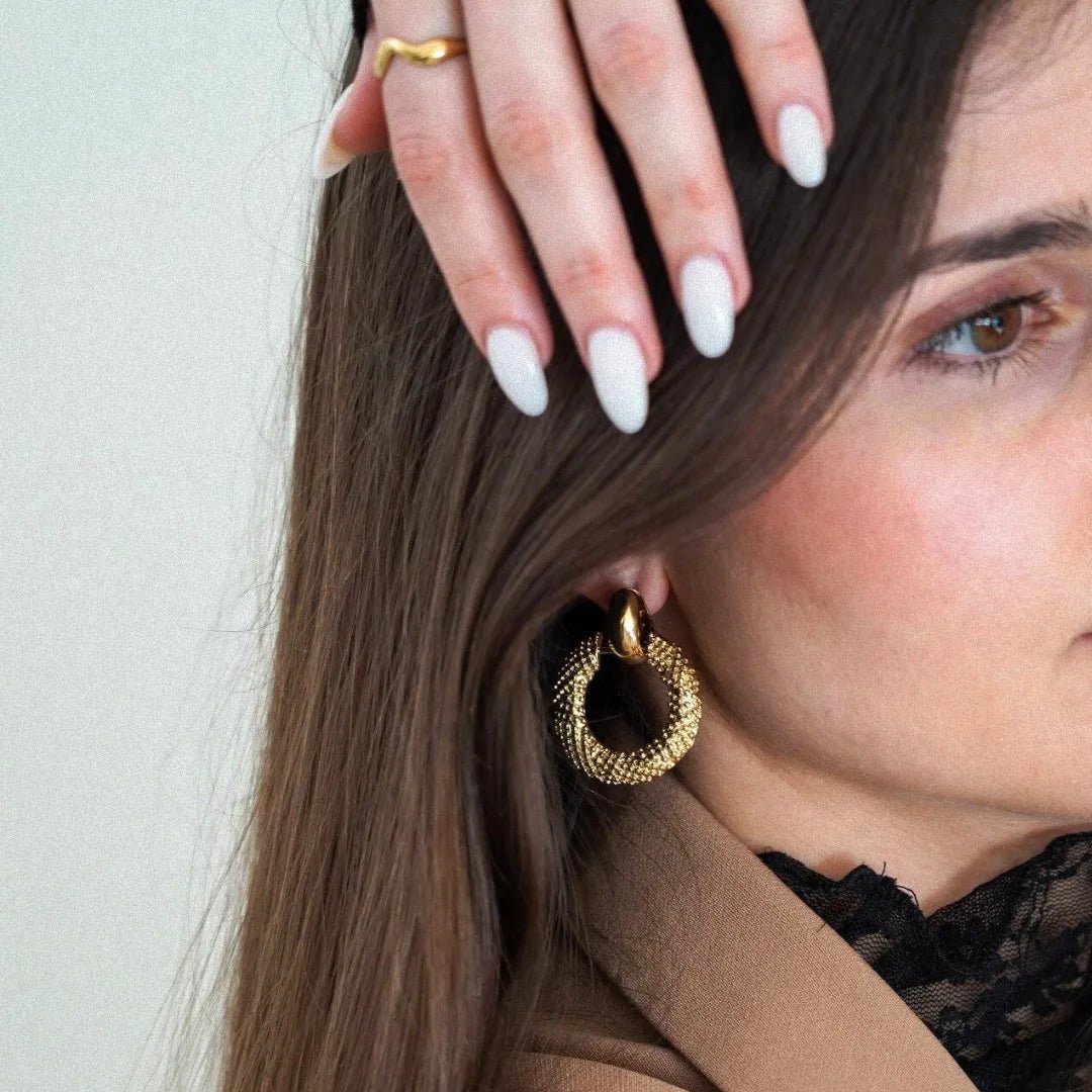 Boucle d'oreille anneau doré à maille tressée, portée par une femme aux cheveux bruns, main avec bague dorée et ongles blancs.