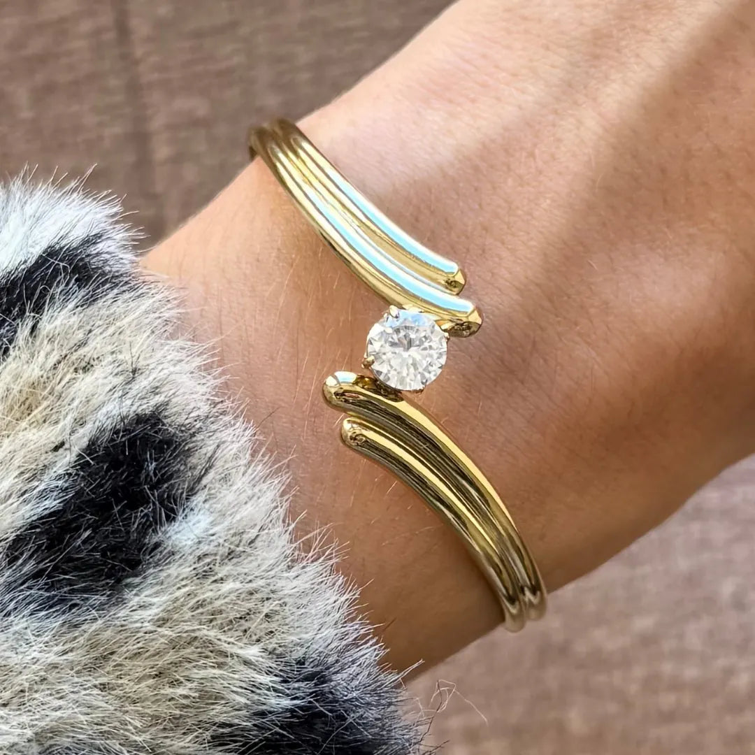 Bracelet jonc ouvert en acier inoxydable doré, croisé multirang avec un zircon solitaire, porté sur un poignet.