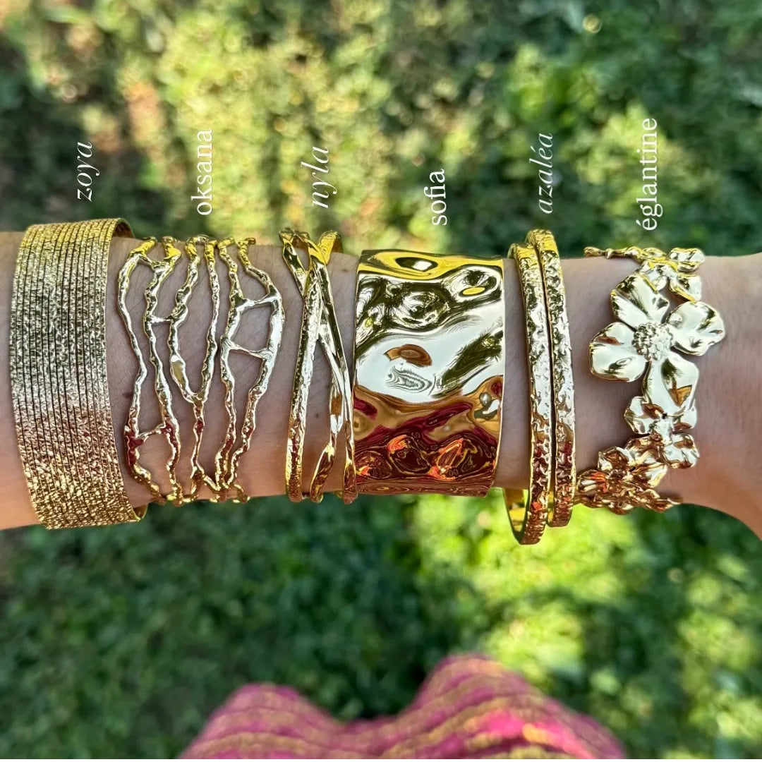 Six bracelets en acier inoxydable doré, aux motifs variés: texturé, organique, martelé et floral, portés sur un poignet.
