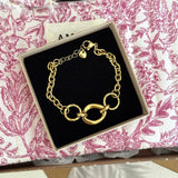 Bracelet doré, maillons ronds texturés et ovale lisse, fermoir mousqueton, breloque cœur, boîte noire sur tissu fleuri rose.