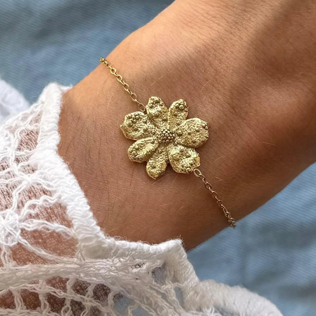 Bracelet fin en acier inoxydable doré, avec pendentif fleur texturé sur un poignet. Manches en dentelle blanche.