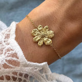 Bracelet fin en acier inoxydable doré, avec pendentif fleur texturé sur un poignet. Manches en dentelle blanche.