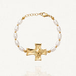 Bracelet doré composé de perles baroques blanches et de petites billes, centré sur une croix texturée avec un cœur rayonnant. Fermoir mousqueton et chaînette d'extension visibles.