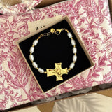 Bracelet doré à perles baroques blanches et croix texturée dorée avec cœur rayonnant, fermoir mousqueton, dans un écrin noir.