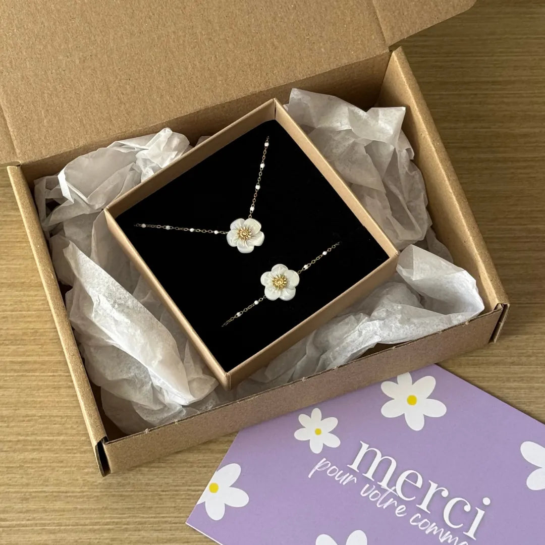 Un coffret cadeau ouvert contient un bracelet et un collier en acier inoxydable doré avec fleurs nacrées, perles et strass.