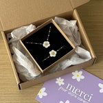 Un coffret cadeau ouvert contient un bracelet et un collier en acier inoxydable doré avec fleurs nacrées, perles et strass.