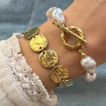 Bracelet doré à disques martelés et bracelet perles baroques blanches avec fermoir toggle or sur poignet.