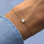 Bracelet fin en acier inoxydable doré, avec un carré en jade blanc, porté au poignet.