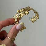Bracelet manchette en acier inoxydable doré, orné de multiples fleurs texturées, tenu par une main.