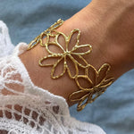 Bracelet jonc en acier inoxydable doré, décoré de trois fleurs ajourées et texturées, porté sur un poignet avec une manche en