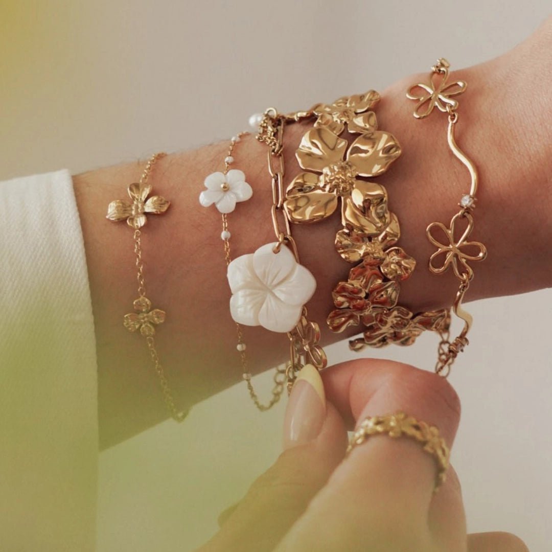 Bracelets en acier inoxydable doré. Motifs floraux variés, avec des fleurs blanches style nacre, superposés sur un poignet.