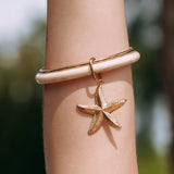 Bracelet jonc en acier inoxydable doré avec partie beige marbrée et charm étoile de mer texturée, sur un bras.