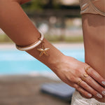 Bracelet jonc en acier inoxydable doré avec charm étoile de mer texturée, sur poignet de femme devant piscine.