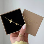 Bracelet fin doré en acier inoxydable avec deux pendentifs fleurs, présenté dans une boîte cadeau noire.