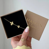 Bracelet fin doré en acier inoxydable avec deux pendentifs fleurs, présenté dans une boîte cadeau noire.