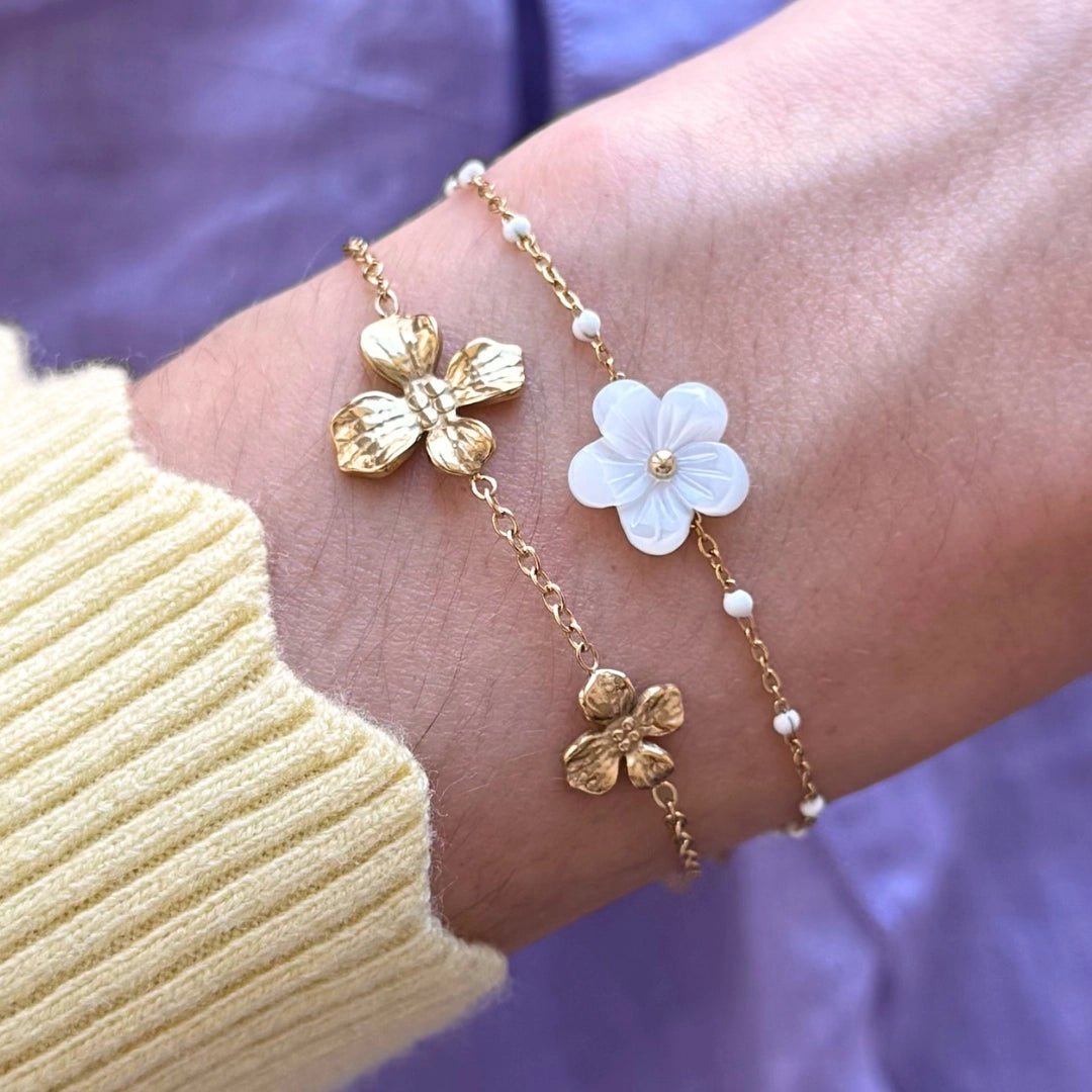 Bracelets en acier inoxydable dorés: un avec fleurs dorées, l'autre avec fleur en nacre et perles blanches sur un poignet.