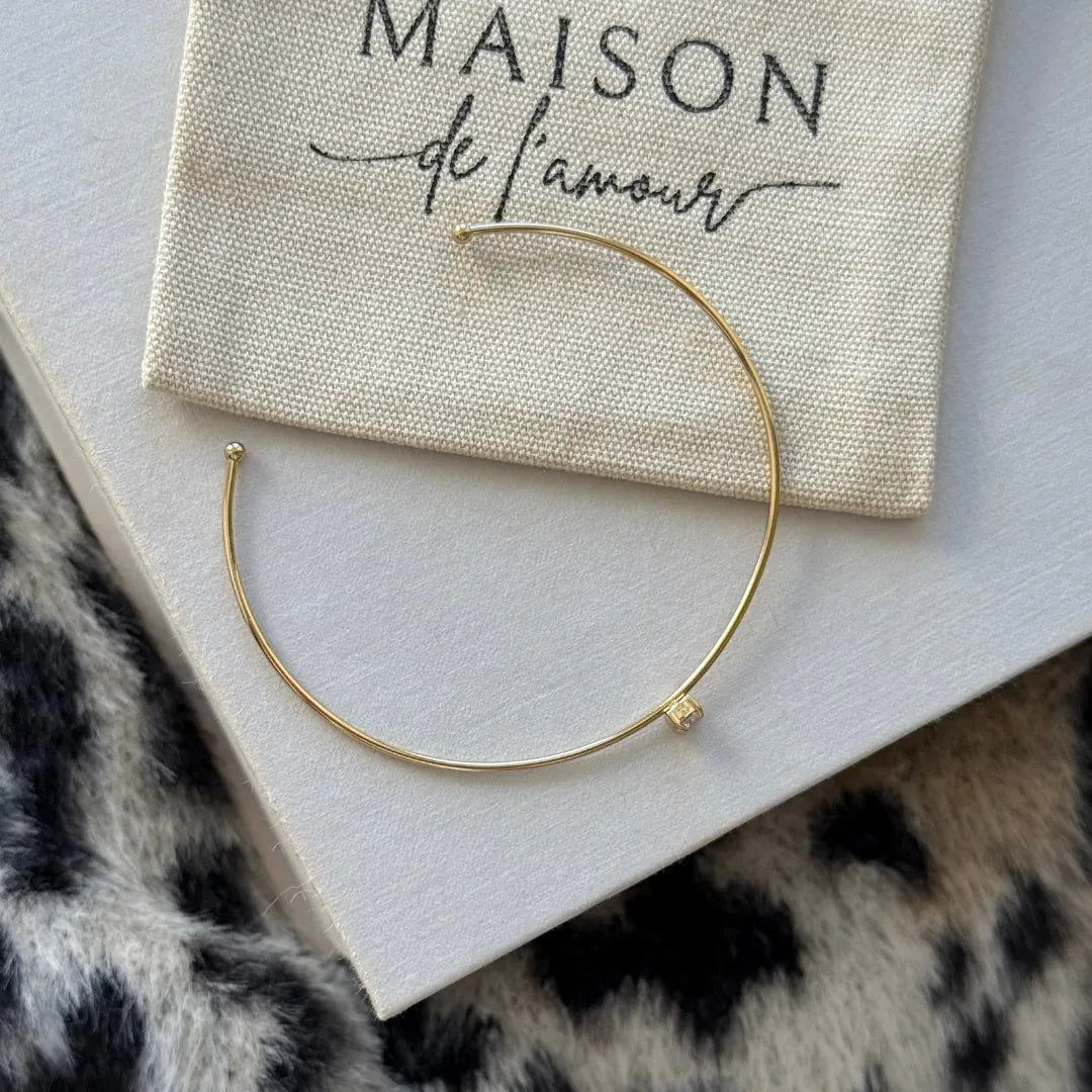 Bracelet jonc fin ouvert en acier inoxydable doré, avec un zircon carré central, posé sur un pochon en tissu blanc et un fond