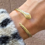 Bracelet jonc serpent ouvert en acier inoxydable doré, à double tête et corps sculptés d'écailles, porté au poignet.