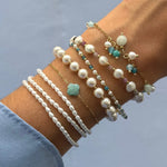 Plusieurs bracelets en acier inoxydable doré portés sur un avant-bras, dont des rangées de perles blanches et un trèfle en