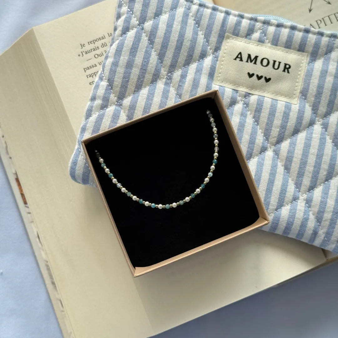 Bracelet acier inoxydable doré, perles blanches et apatite bleue, présenté dans sa boîte, près d'un livre et d'une pochette