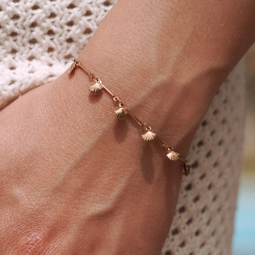 Bracelet fin en acier inoxydable doré, orné de multiples petites coquilles, porté au poignet.