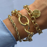 Trois bracelets en acier inoxydable doré sur un poignet: tressé, jonc maximaliste nœud, et chaîne à breloque martelée.