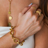Bracelet chaîne à grosses mailles en acier inoxydable doré, porté au poignet d'une femme avec d'autres bijoux dorés.