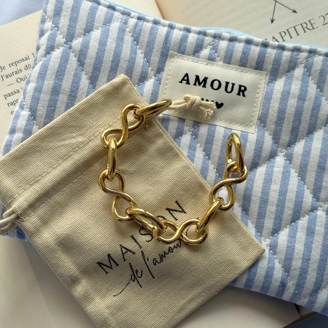 Bracelet en acier inoxydable doré à maillons en forme de nœuds, posé sur un pochon beige et un sac rayé bleu et blanc.