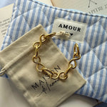 Bracelet en acier inoxydable doré à maillons en forme de nœuds, posé sur un pochon beige et un sac rayé bleu et blanc.