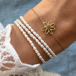 Un poignet porte un bracelet acier inoxydable doré à fleur ajourée et un à trois rangs de perles blanches.