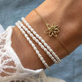 Un poignet porte un bracelet acier inoxydable doré à fleur ajourée et un à trois rangs de perles blanches.
