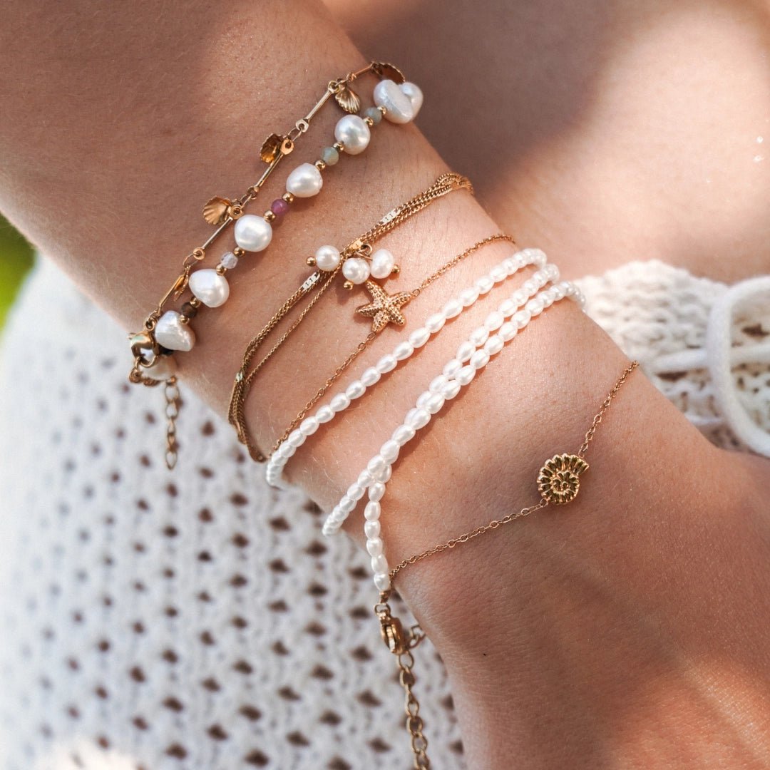 Lot de bracelets en acier inoxydable doré avec perles, coquillages et étoile de mer sur poignet féminin.
