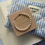 Bracelet perles blanches et acier inoxydable doré sur une boîte rose pâle. Un pochon bleu et blanc est en arrière-plan.