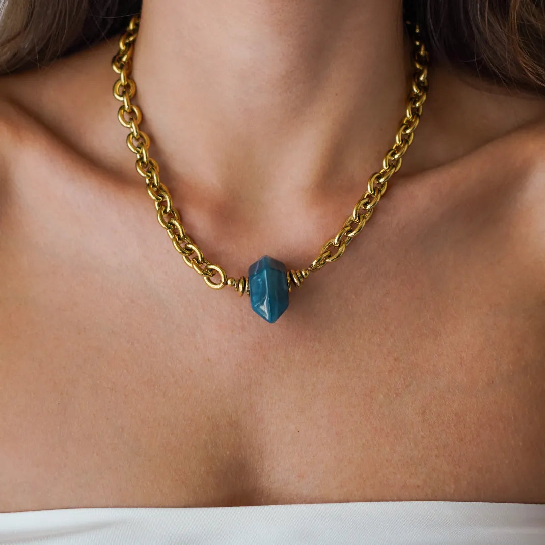 Collier en acier inoxydable doré à maille épaisse, orné d'un pendentif pierre bleue en acétate, porté au cou.