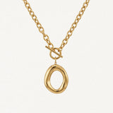 Collier BENEDICTE en acier inoxydable doré. Chaîne à maillons ovales, fermoir toggle et grand pendentif ovale de forme organique irrégulière. Posé sur fond blanc.