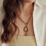 Collier chaîne dorée à maillons ovales, fermoir toggle et pendentif anneau ovale irrégulier, porté sur décolleté de femme.