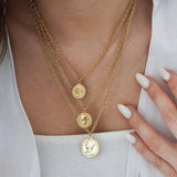 Trois colliers dorés superposés, pendentifs pièces de monnaie buste profil de tailles diverses, sur femme, main ongles blancs.