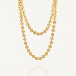 Collier long doré à deux rangs composé de multiples disques ronds martelés en acier inoxydable, posé sur un fond blanc avec un léger reflet.