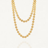 Collier long doré à deux rangs composé de multiples disques ronds martelés en acier inoxydable, posé sur un fond blanc avec un léger reflet.
