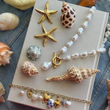 Collier de perles baroques blanches avec fermoir T en acier inoxydable doré, sur un livre blanc entouré de coquillages et