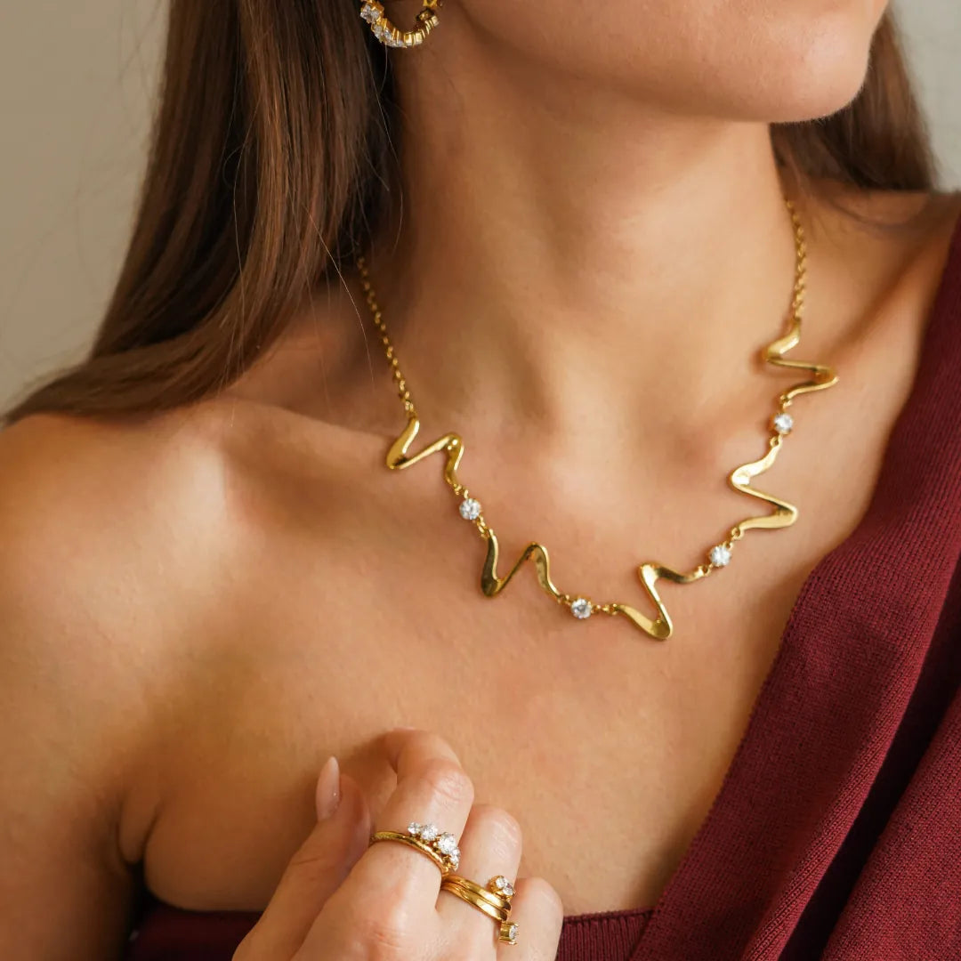 Collier ondulé en acier inoxydable doré, orné de six zircons ronds, porté sur un décolleté.