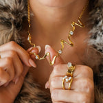 Collier ondulé en acier inoxydable doré orné de zircons ronds. Une femme le tient devant son cou. Elle porte une bague