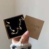 Collier ondulé en acier inoxydable doré et zircons, dans une boîte cadeau marron ouverte tenue par une main.