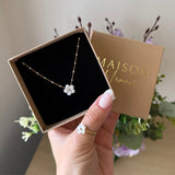 Collier maille perlée en acier inoxydable doré avec pendentif fleur en nacre blanche, présenté dans un écrin marron. Une