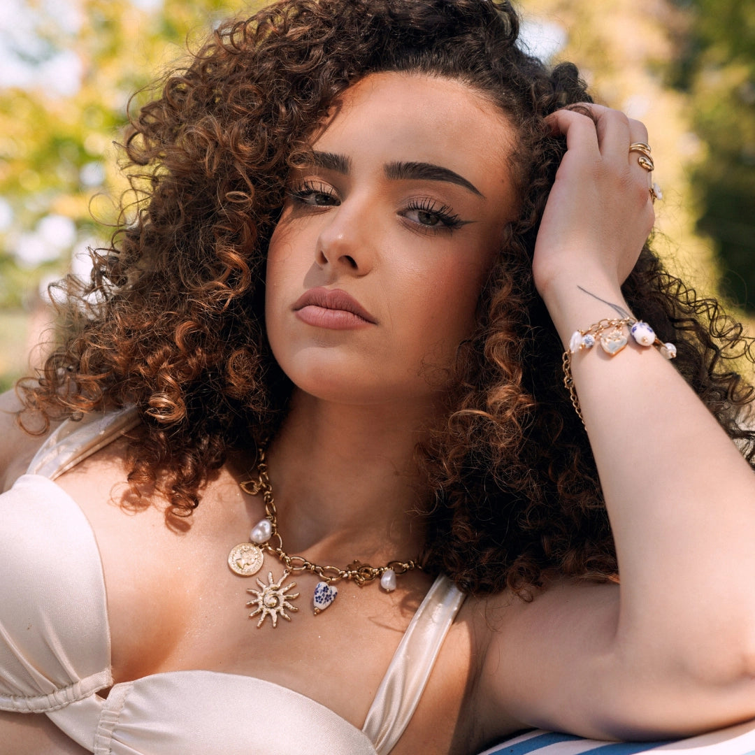 Collier en acier inoxydable doré avec pendentifs soleil, pièce, cœur bleu et perles, porté par une femme.