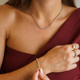 Une femme au haut bordeaux porte un collier rivière tennis en acier inoxydable doré serti de zircons et un bracelet doré.