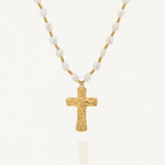 Collier avec une chaîne composée de perles blanches baroques et de petites billes dorées. Le pendentif est une croix dorée à la surface texturée et irrégulière.